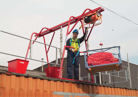 A construction worker wearing a helmet, safety harness and hi-vis vest operates a red gantry hoist
