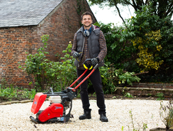Man standing on a gravel surface operating a red plate compactor in a garden area