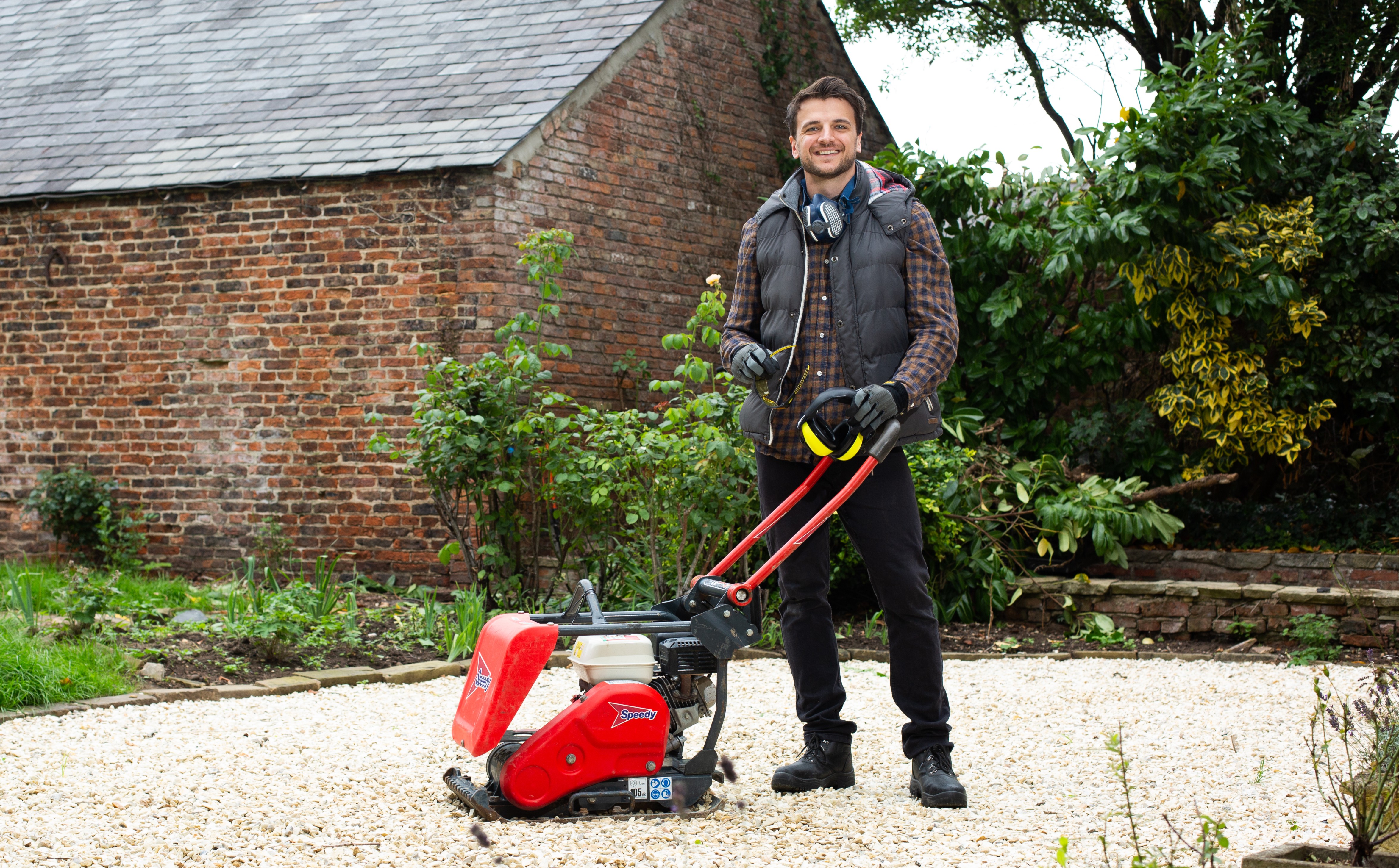 Man standing on a gravel surface operating a red plate compactor in a garden area