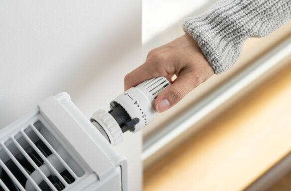 A hand turns the temperature dial on a white radiator thermostat attached to a wall-mounted heater.