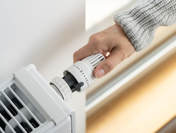 A hand turns the temperature dial on a white radiator thermostat attached to a wall-mounted heater.