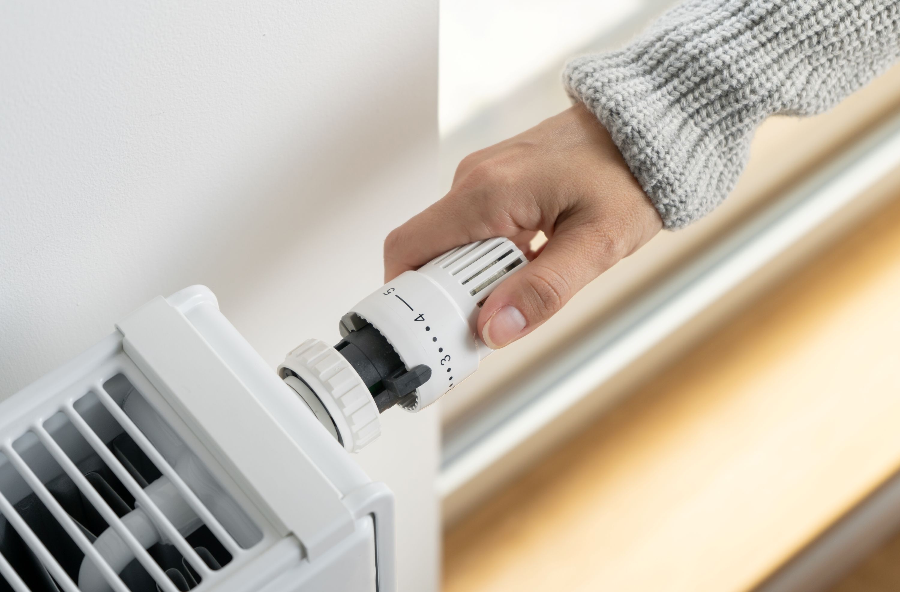 A hand turns the temperature dial on a white radiator thermostat attached to a wall-mounted heater.