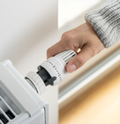 A hand turns the temperature dial on a white radiator thermostat attached to a wall-mounted heater.