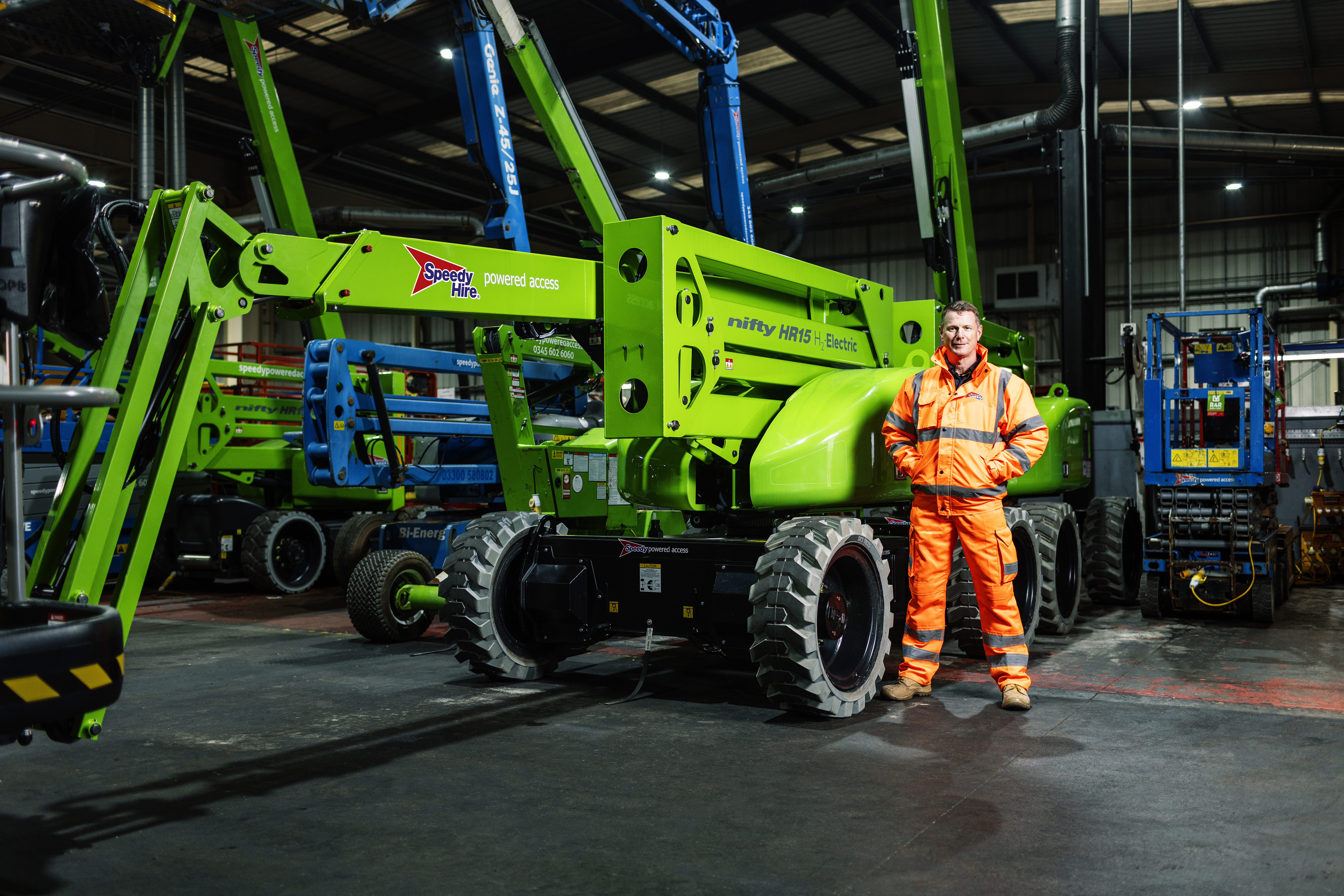 Speedy Hire Nifty HR15 H2 Electric Nifty Lift. Green powered access nifty lift with employee in highvis clothing stood to the right. 