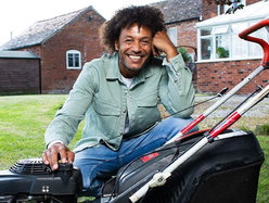 Person sitting on grass next to a black and red lawn mower in front of a brick house