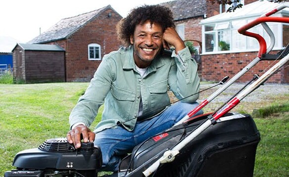 Person sitting on grass next to a black and red lawn mower in front of a brick house