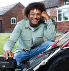 Person sitting on grass next to a black and red lawn mower in front of a brick house