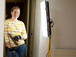 Woman standing in a doorway holding a trowel, illuminated by a bright work light during a DIY project.