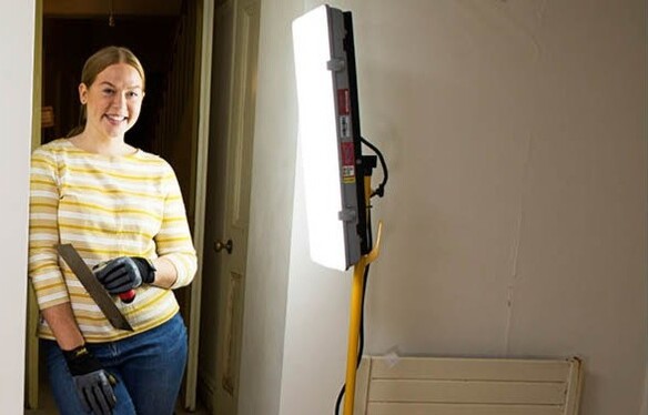 Woman standing in a doorway holding a trowel, illuminated by a bright work light during a DIY project.