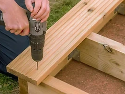 Person using a power drill to assemble a wooden frame outdoors.