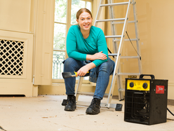 A portable industrial fan heater placed on the floor of a room under renovation, with a person seated on a ladder in the background holding a tool.
