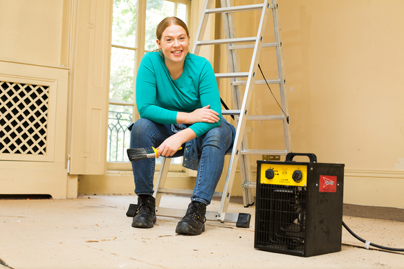 A portable industrial fan heater placed on the floor of a room under renovation, with a person seated on a ladder in the background holding a tool.