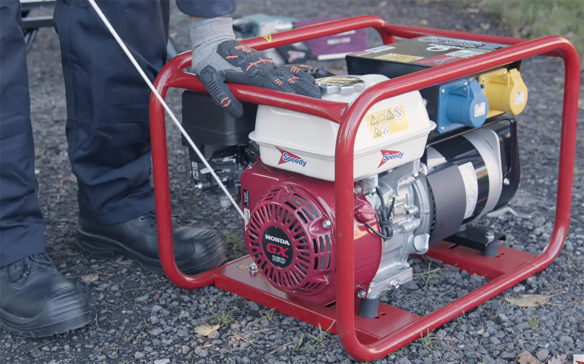 A person wearing protective gloves pulls the start cord on a portable petrol generator with a red frame and Honda GX engine.