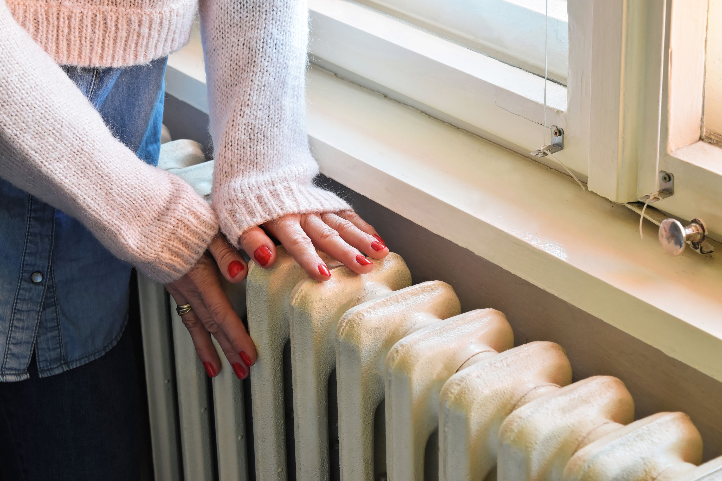 Close-up of a person&rsquo;s hands resting on a white radiator beneath a window.