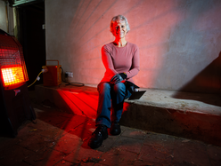 Woman sitting indoors near a glowing red portable heater, wearing gloves and casual work clothes.