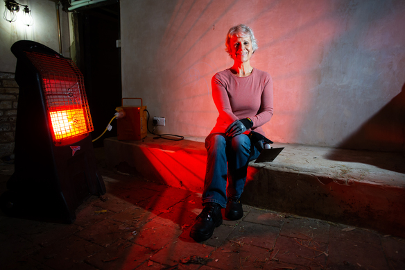 Woman sitting indoors near a glowing red portable heater, wearing gloves and casual work clothes.
