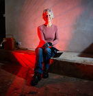 Woman sitting indoors near a glowing red portable heater, wearing gloves and casual work clothes.