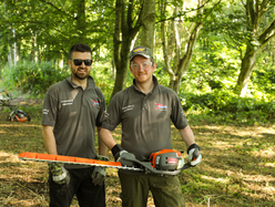 Two Speedy staff members holding a hedge trimmer while standing in a wooded area.
