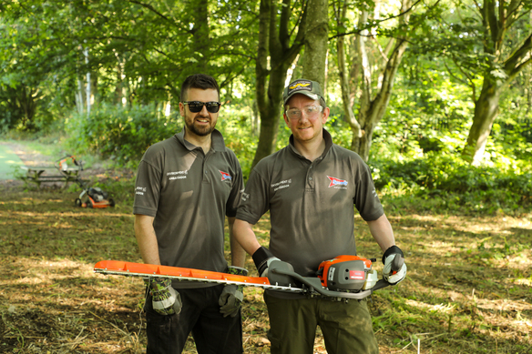 Two Speedy staff members holding a hedge trimmer while standing in a wooded area.