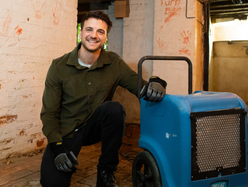 Person kneeling beside a blue portable dehumidifier in a basement with white brick walls, exposed pipes, and a single light bulb hanging from the ceiling.