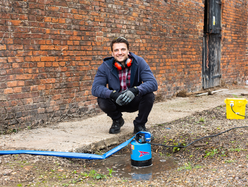 Person using a water pump to drain standing water near a brick wall.