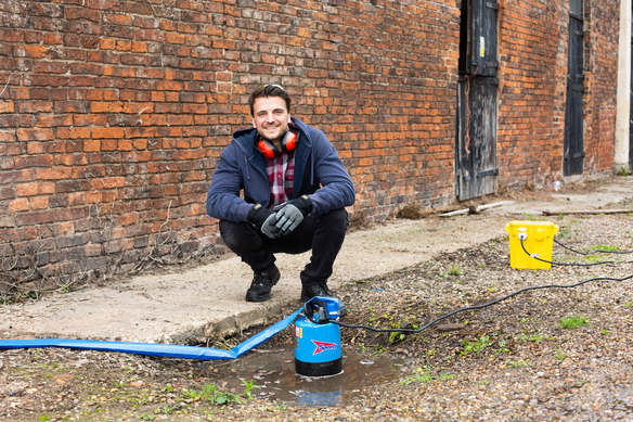 Person using a water pump to drain standing water near a brick wall.