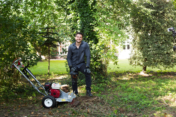 Person using a stump grinder to remove tree roots in a garden area.