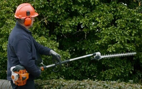 Person using a long-reach hedge trimmer to trim tall garden hedges.