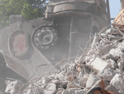 Close-up of heavy demolition equipment crushing a pile of concrete rubble, with dust rising in the air and green trees visible in the background.