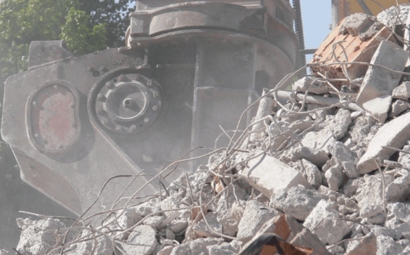 Close-up of heavy demolition equipment crushing a pile of concrete rubble, with dust rising in the air and green trees visible in the background.