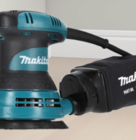 Makita detail sander placed on a light wooden floor with dust bag attached.