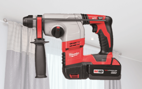 Red Milwaukee M18 cordless rotary hammer drill displayed in a bright room with curtains and ceiling spotlights.