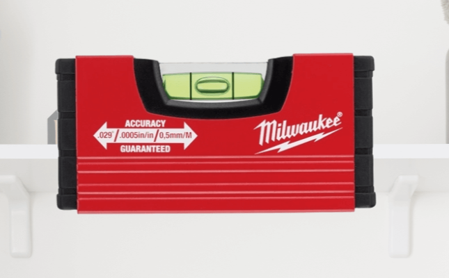 Red Milwaukee spirit level placed on a white wall shelf with decorative items in the background.