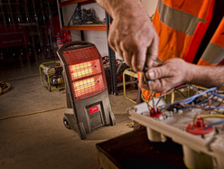 A portable infrared heater glows red in a workshop while a worker in a high‑visibility vest adjusts electrical components on a workbench.