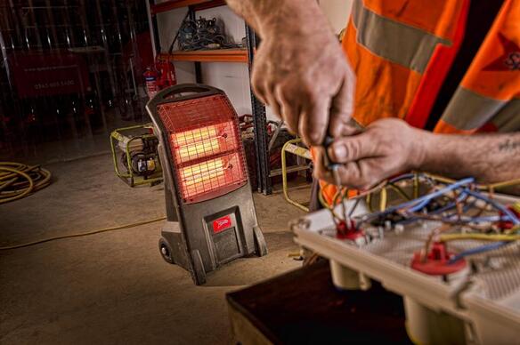 A portable infrared heater glows red in a workshop while a worker in a high‑visibility vest adjusts electrical components on a workbench.