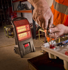 A portable infrared heater glows red in a workshop while a worker in a high‑visibility vest adjusts electrical components on a workbench.