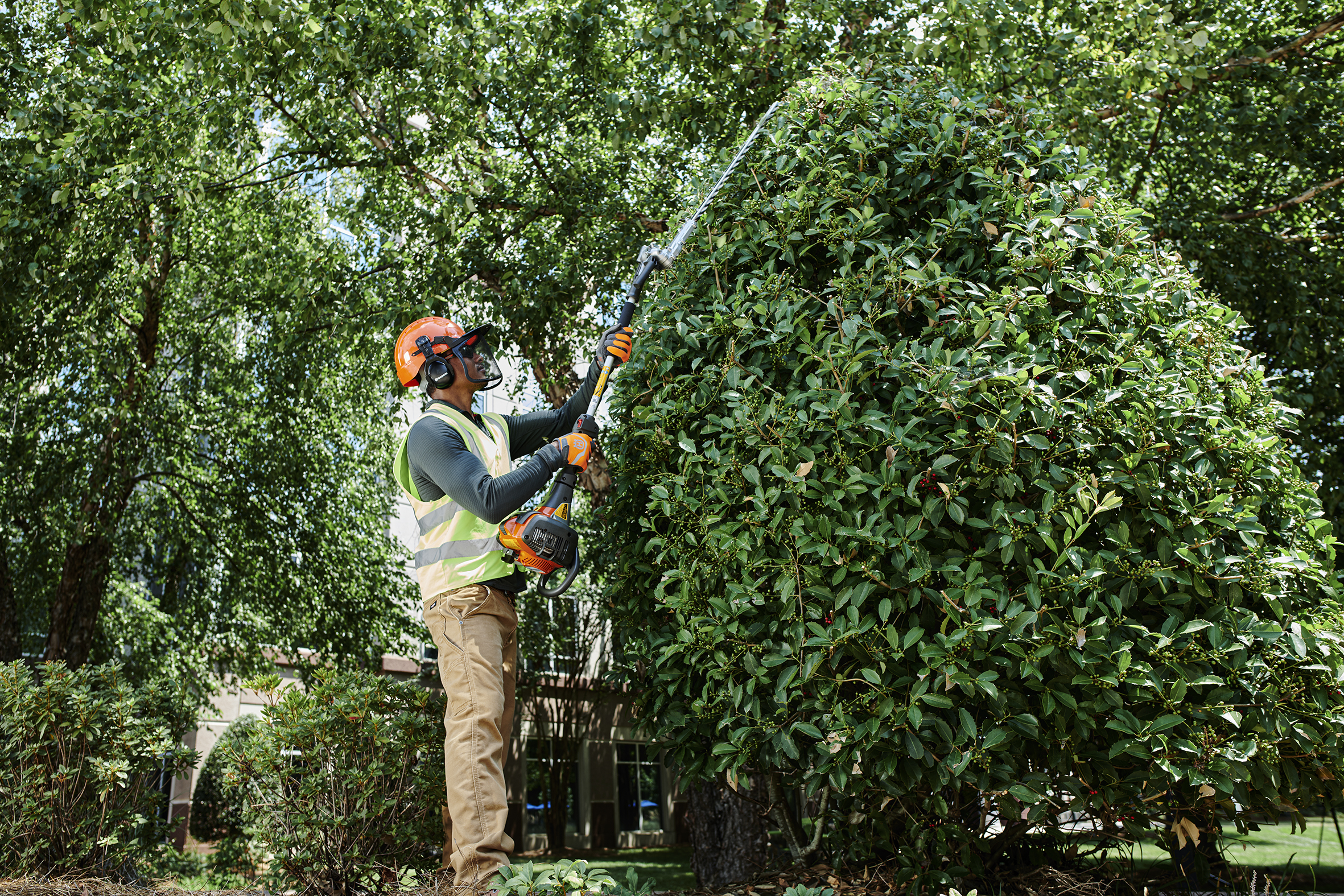 Worker using a long‑reach hedge trimmer to shape a tall garden shrub, wearing safety gear and a high‑visibility vest.