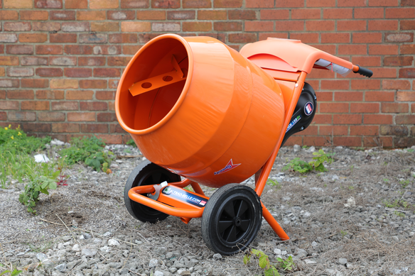 Bright orange portable cement mixer with black wheels placed on gravel near a brick wall.