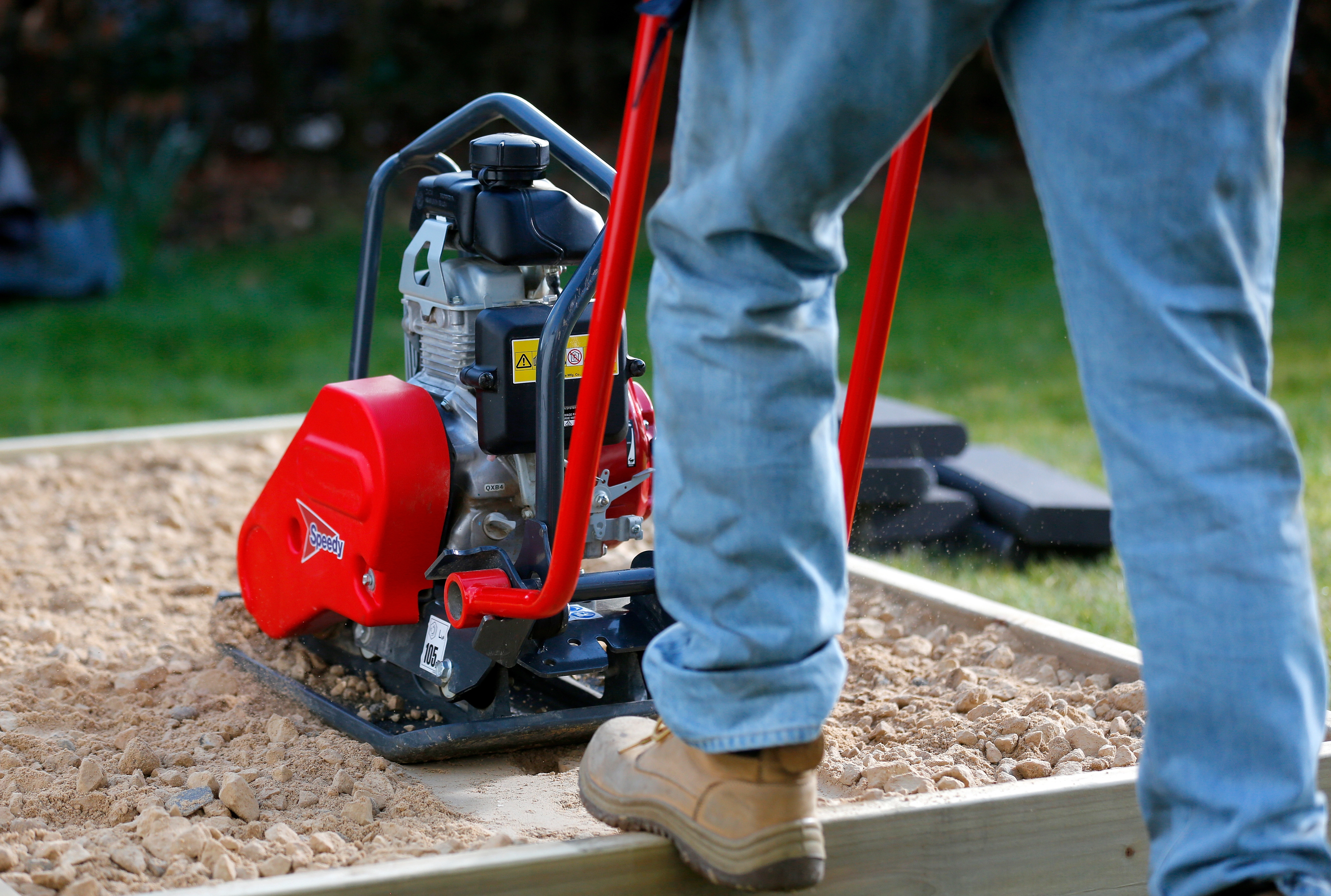 Person operating a red Speedy plate compactor on a sand and gravel surface.