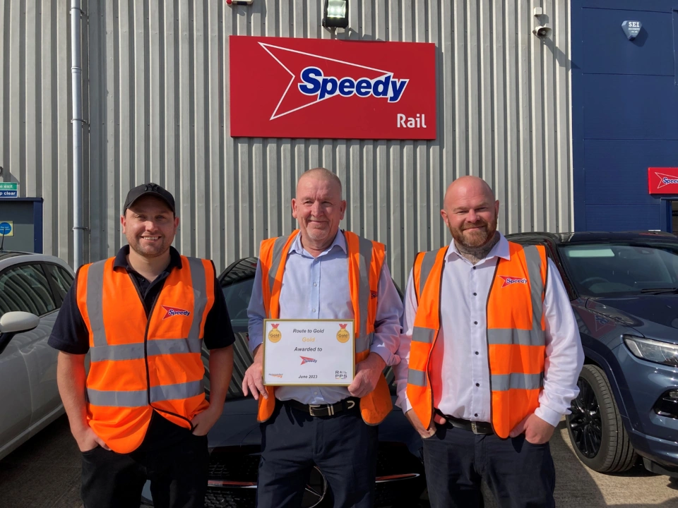 Three men in orange safety vests standing outside a Speedy Rail depot, with the man in the centre holding a gold award certificate.