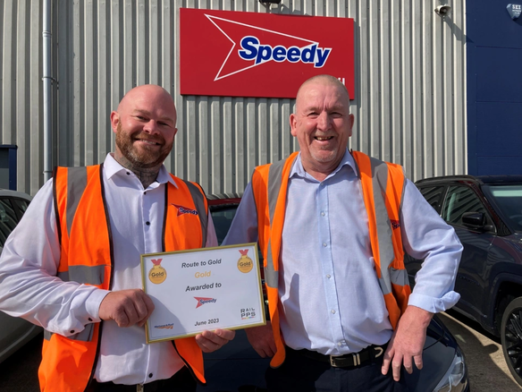 Two men wearing orange safety vests smiling outside a Speedy depot, holding a framed gold award certificate.