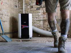 Person in work boots and shorts walking inside a room under renovation with an air conditioning unit labeled 'Speedy'