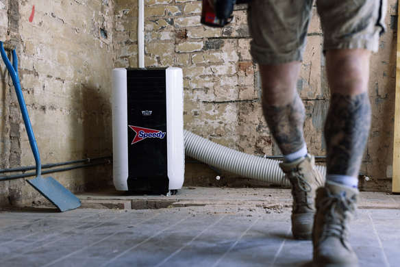Person in work boots and shorts walking inside a room under renovation with an air conditioning unit labeled 'Speedy'