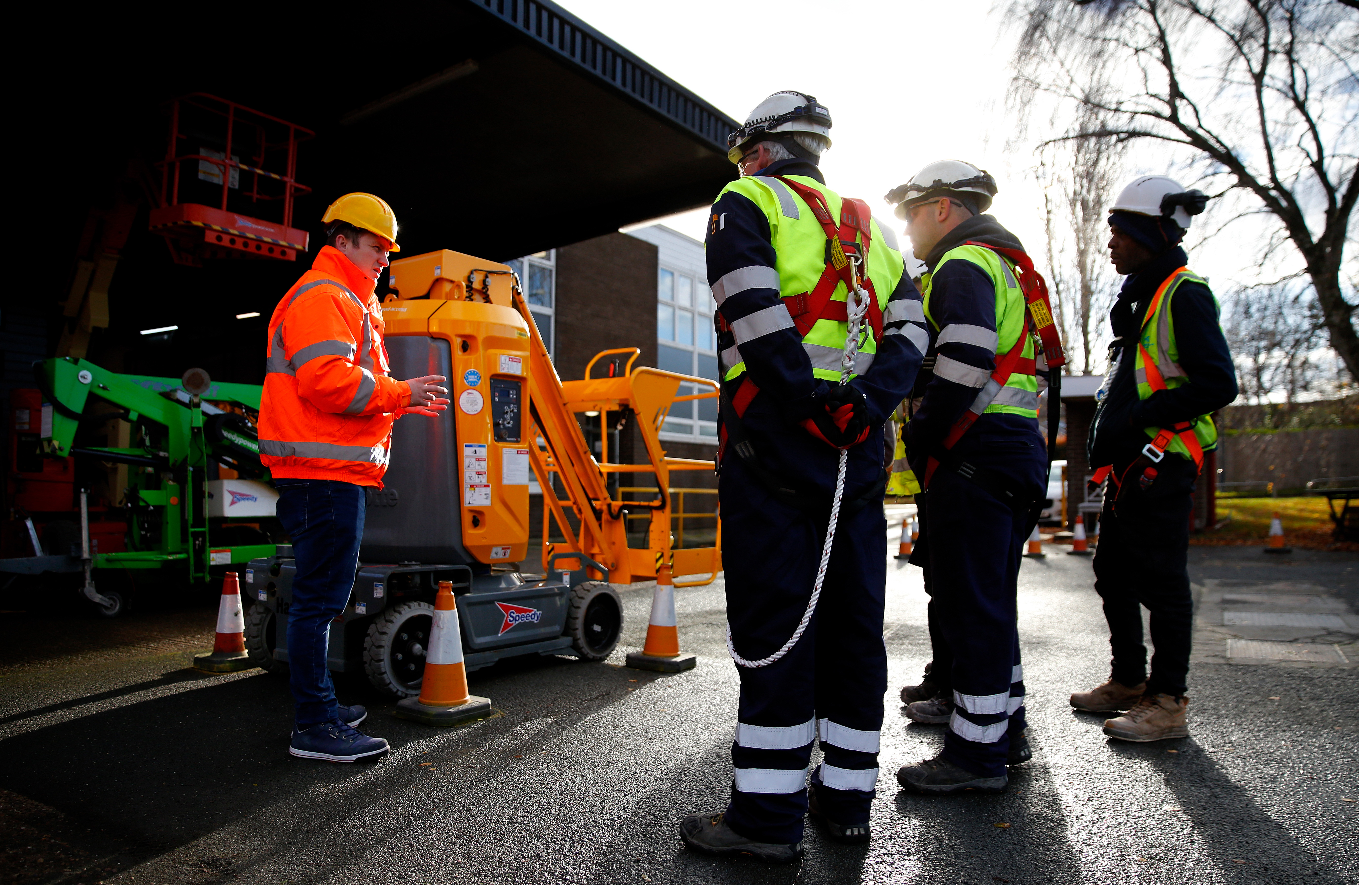 Instructor demonstrating a mobile elevating work platform to workers wearing safety harnesses and high‑visibility gear.