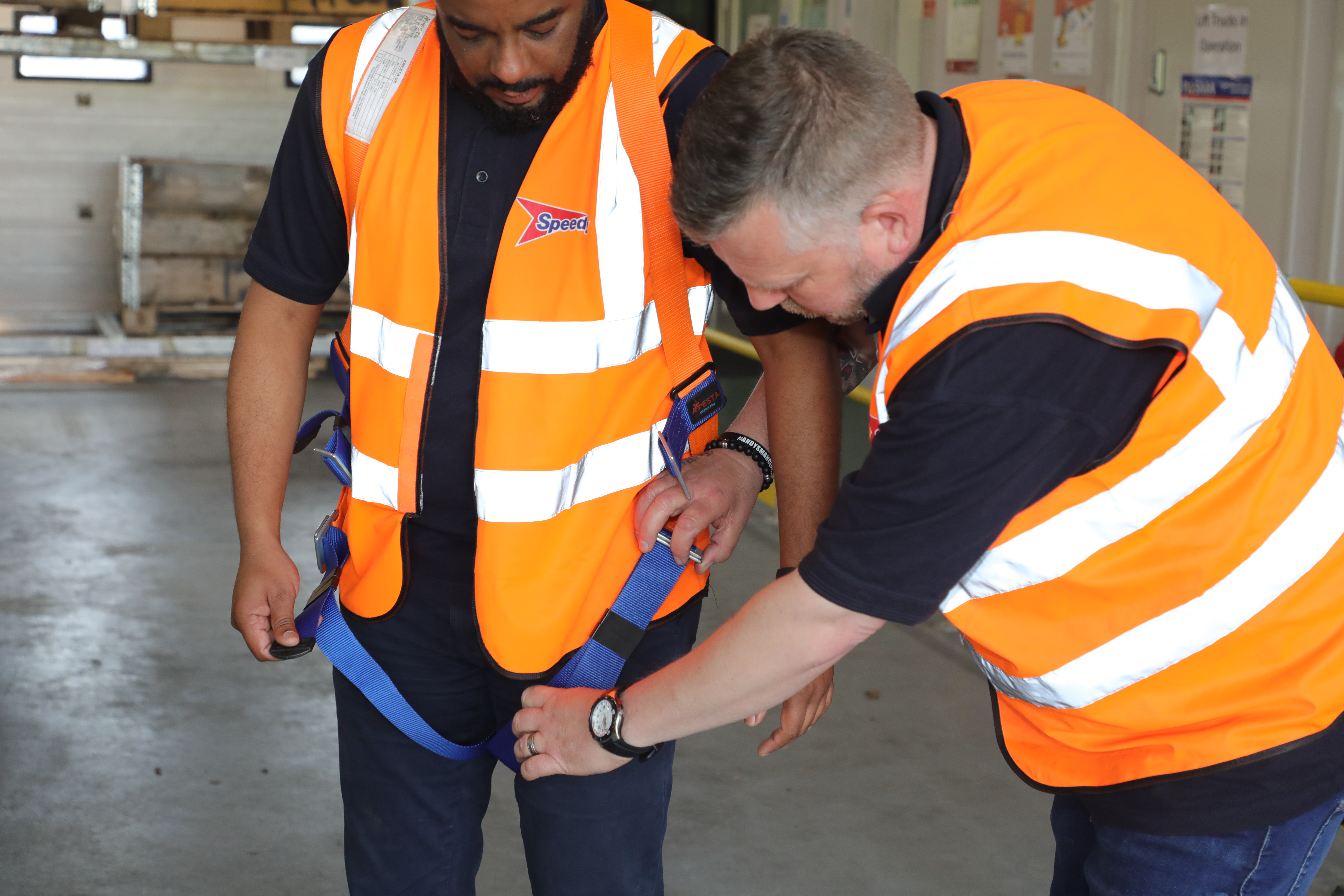 Two workers in orange high‑visibility vests fitting and adjusting a safety harness.
