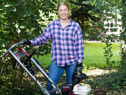Person standing outdoors in a garden area holding a petrol-powered turf cutter, surrounded by green grass, trees, and shrubs
