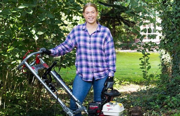 Person standing outdoors in a garden area holding a petrol-powered turf cutter, surrounded by green grass, trees, and shrubs