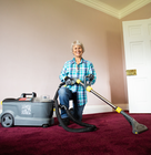 A person uses a professional carpet extraction cleaner on a deep red carpet in an empty room, operating the hose and floor wand attachment.