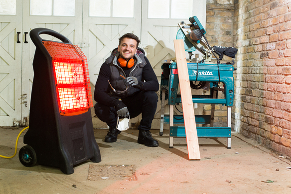 A portable infrared heater glowing red in a workshop, positioned near a worker and woodworking tools on the floor.