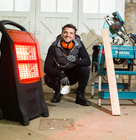 A portable infrared heater glowing red in a workshop, positioned near a worker and woodworking tools on the floor.